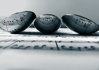 Three transparent marbles sit atop a sheet of music, with the musical notes clearly visible through the marbles. The arrangement creates an artistic distortion of the musical notes, adding a unique visual effect. The focus is soft, enhancing a sense of calmness.