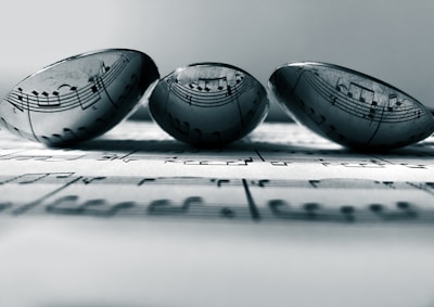 Three transparent marbles sit atop a sheet of music, with the musical notes clearly visible through the marbles. The arrangement creates an artistic distortion of the musical notes, adding a unique visual effect. The focus is soft, enhancing a sense of calmness.