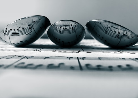 Three transparent marbles sit atop a sheet of music, with the musical notes clearly visible through the marbles. The arrangement creates an artistic distortion of the musical notes, adding a unique visual effect. The focus is soft, enhancing a sense of calmness.