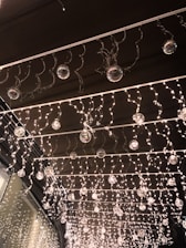 A decorative installation featuring numerous small twinkling lights hanging from a ceiling. Glass orbs are interspersed throughout the lights, creating a shimmering effect. The lights and orbs are organized in neat rows, presenting a festive and elegant atmosphere.