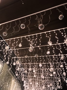 A decorative installation featuring numerous small twinkling lights hanging from a ceiling. Glass orbs are interspersed throughout the lights, creating a shimmering effect. The lights and orbs are organized in neat rows, presenting a festive and elegant atmosphere.