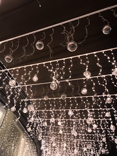 A decorative installation featuring numerous small twinkling lights hanging from a ceiling. Glass orbs are interspersed throughout the lights, creating a shimmering effect. The lights and orbs are organized in neat rows, presenting a festive and elegant atmosphere.