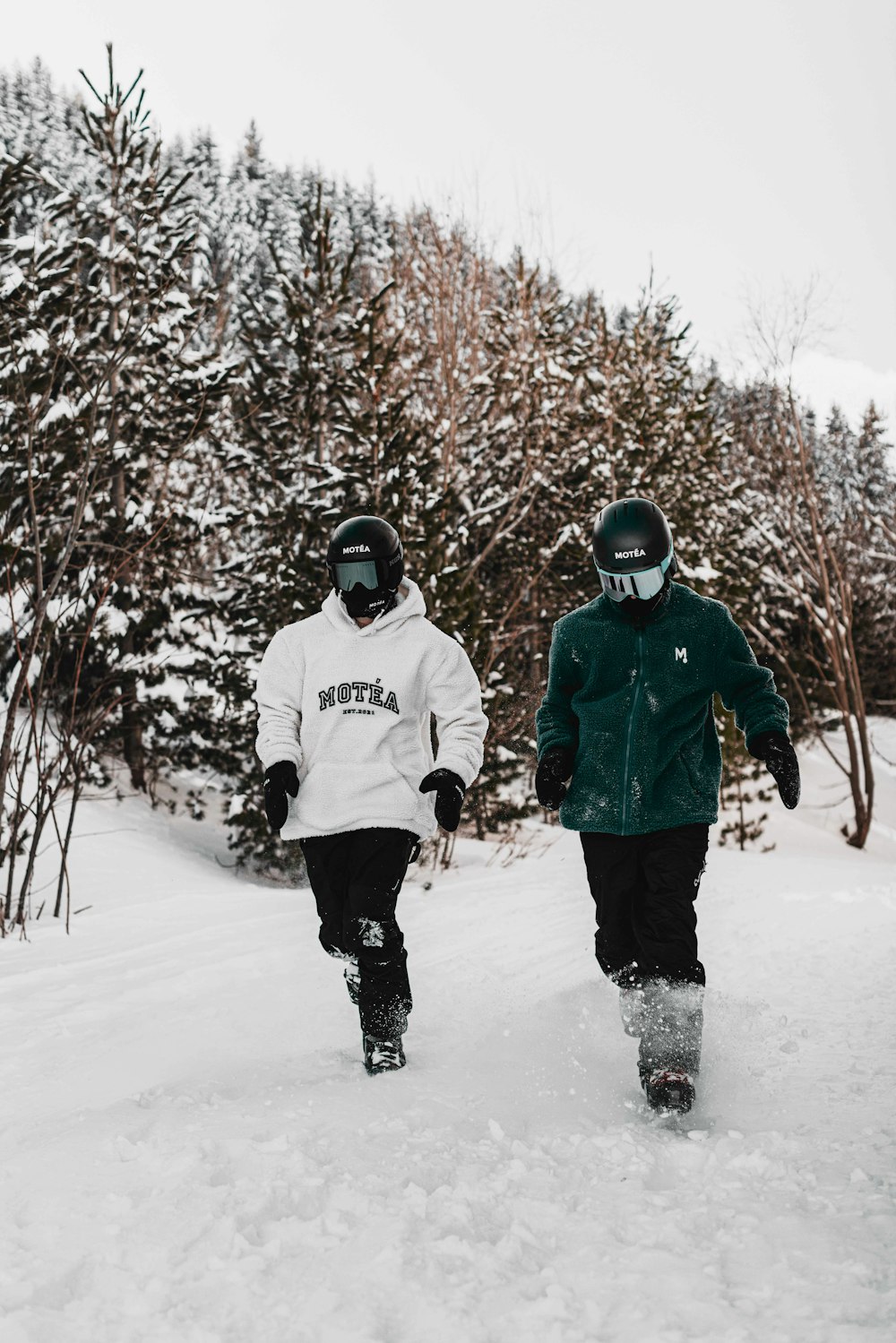 two snowboarders walking through the snow in front of trees