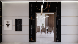 A modern interior features a sleek, minimalist design with a large doorway leading into a dining area. The walls are adorned with black and white marble details, complemented by geometric artwork. The dining room has a wooden table surrounded by upholstered chairs, and a contemporary chandelier hangs overhead.