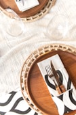 A neatly arranged table setting featuring woven placemats, wooden plates, and black-and-white patterned napkins. Cutlery, consisting of a fork and knife, rests on the napkin. Two empty wine glasses are placed near the setting on a textured white tablecloth.