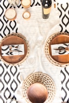 A table setting featuring two woven placemats, each with a wooden plate and a napkin with a black and white pattern. A bottle of wine and three candles are arranged on a white textured table runner over a black and white geometric patterned tablecloth.