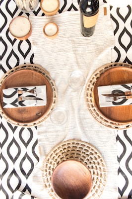 A table setting featuring two woven placemats, each with a wooden plate and a napkin with a black and white pattern. A bottle of wine and three candles are arranged on a white textured table runner over a black and white geometric patterned tablecloth.