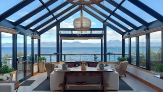 A spacious and modern sunroom with large glass windows and a glass ceiling, overlooking a scenic view of the sea and distant mountains. The room is elegantly furnished with comfortable sofas, armchairs, and a wooden table arranged with decorative items. Potted plants add a touch of greenery to the space, and a woven pendant lamp hangs from the ceiling, enhancing the room's warmth and ambiance.