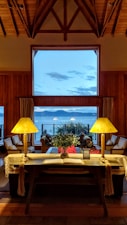 A warmly lit living room with high wooden ceilings and a large window overlooking the sea. In the foreground, there is a table with two lamps, an open book, and some greenery. Comfortable chairs and sofas are arranged facing the window, providing a cozy and inviting atmosphere.