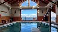 A serene indoor pool area framed with wooden beams and large windows providing a view of a distant mountain across a body of water. Natural light fills the space, with poolside seating available. A lifebuoy is mounted on the wall, and decorative tall vases are positioned against the walls.