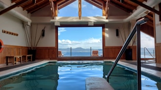 A serene indoor pool area framed with wooden beams and large windows providing a view of a distant mountain across a body of water. Natural light fills the space, with poolside seating available. A lifebuoy is mounted on the wall, and decorative tall vases are positioned against the walls.
