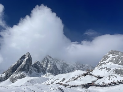 Majestic snow-covered mountains rise against a deep blue sky, partially obscured by thick white clouds. A wooden walkway winds up the slope, with small groups of people visible making their way along it.