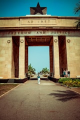 A large archway monument constructed of beige stone with the inscription 'AD 1957' and 'FREEDOM AND JUSTICE'. The top of the monument features a prominent black star. A person walks along the pathway leading to the arch, flanked by neatly lined palm trees. There are a few people standing on the right side, near the base of the monument.
