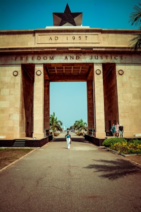 A large archway monument constructed of beige stone with the inscription 'AD 1957' and 'FREEDOM AND JUSTICE'. The top of the monument features a prominent black star. A person walks along the pathway leading to the arch, flanked by neatly lined palm trees. There are a few people standing on the right side, near the base of the monument.