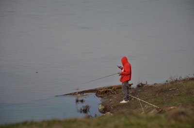 A person wearing a red hoodie stands near the edge of a calm body of water, holding a fishing rod while looking at a mobile phone. The ground around is grassy and slightly muddy with a few rocks and sparse vegetation.