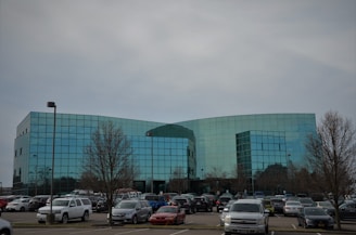 A modern, glass-fronted office building with a curved structure is situated in the middle of a spacious parking lot filled with various cars. Trees without leaves are scattered around the lot, and the sky is overcast.