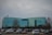 A modern, glass-fronted office building with a curved structure is situated in the middle of a spacious parking lot filled with various cars. Trees without leaves are scattered around the lot, and the sky is overcast.