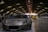 A sleek sports car with a metallic finish is prominently displayed in an indoor venue with arched ceilings and dim lighting. Several other vehicles are lined up in the background, including a bright yellow car. People are gathered around, some observing the vehicles.