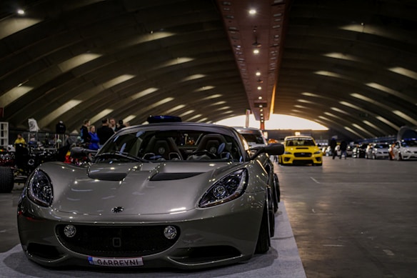 A sleek sports car with a metallic finish is prominently displayed in an indoor venue with arched ceilings and dim lighting. Several other vehicles are lined up in the background, including a bright yellow car. People are gathered around, some observing the vehicles.