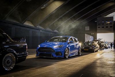 A row of cars is parked inside a large, dimly lit garage with an arched ceiling. The cars vary in type and color, with a prominent blue sports car in the foreground. Light beams filter through the air, creating shadows and highlighting the vehicles.
