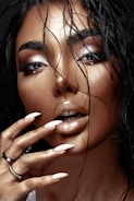 A close-up image of a person with wet hair cascading over their face, accentuating their flawless makeup and glossy lips. Long fingernails, painted in a nude color, touch the face softly while a few subtle jewelry pieces are visible on the hand.
