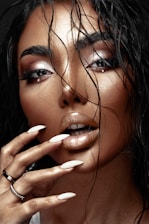 A close-up image of a person with wet hair cascading over their face, accentuating their flawless makeup and glossy lips. Long fingernails, painted in a nude color, touch the face softly while a few subtle jewelry pieces are visible on the hand.