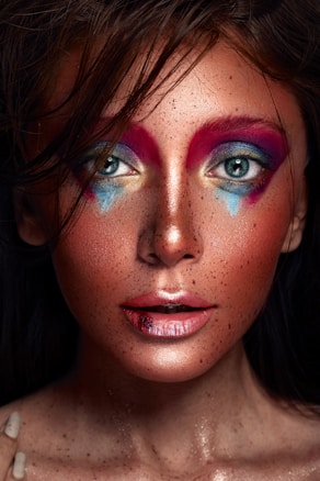 A close-up portrait of a person with elaborate and colorful makeup featuring prominent red and blue eyeshadow extending beyond the eyelids, combined with a glittery finish. The skin is speckled, and the lips are glossy with a hint of a reddish hue. The hair is dark and slightly tousled, adding a dramatic effect.