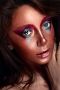 A close-up of a person's face with artistic makeup. Bold, colorful eye makeup features shades of pink, purple, blue, and gold, while the skin glows with shimmering highlights. Freckles are visible across the cheeks and nose. The lips are softly parted with a hint of gloss.