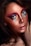 A close-up of a person's face with artistic makeup. Bold, colorful eye makeup features shades of pink, purple, blue, and gold, while the skin glows with shimmering highlights. Freckles are visible across the cheeks and nose. The lips are softly parted with a hint of gloss.