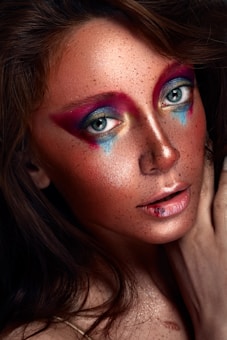 A close-up of a person's face with artistic makeup. Bold, colorful eye makeup features shades of pink, purple, blue, and gold, while the skin glows with shimmering highlights. Freckles are visible across the cheeks and nose. The lips are softly parted with a hint of gloss.