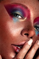 A close-up view of a person's face with vibrant, colorful makeup. The makeup includes shades of pink, blue, and gold around the eyes. The skin has a freckled appearance, and the person is touching their lips with their fingers, which are painted with a neutral polish.