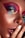 A close-up view of a person's face with vibrant, colorful makeup. The makeup includes shades of pink, blue, and gold around the eyes. The skin has a freckled appearance, and the person is touching their lips with their fingers, which are painted with a neutral polish.