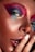 A close-up view of a person's face with vibrant, colorful makeup. The makeup includes shades of pink, blue, and gold around the eyes. The skin has a freckled appearance, and the person is touching their lips with their fingers, which are painted with a neutral polish.