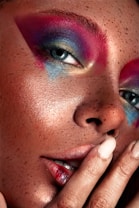 A close-up view of a person's face with vibrant, colorful makeup. The makeup includes shades of pink, blue, and gold around the eyes. The skin has a freckled appearance, and the person is touching their lips with their fingers, which are painted with a neutral polish.