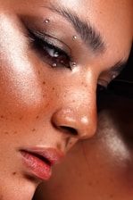 A close-up of a person's face highlighting flawless, dewy skin with makeup. The eye is adorned with dramatic black eyeliner and multiple small, metallic piercings near the eyebrow and eye area. Freckles are visible across the nose and cheeks, adding character.