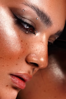 A close-up of a person's face highlighting flawless, dewy skin with makeup. The eye is adorned with dramatic black eyeliner and multiple small, metallic piercings near the eyebrow and eye area. Freckles are visible across the nose and cheeks, adding character.