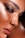 A close-up of a person's face highlighting flawless, dewy skin with makeup. The eye is adorned with dramatic black eyeliner and multiple small, metallic piercings near the eyebrow and eye area. Freckles are visible across the nose and cheeks, adding character.