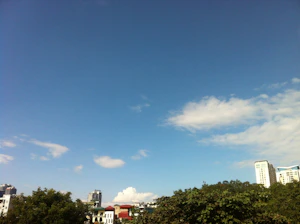 A clear blue sky with sun and light clouds over a bustling urban area.