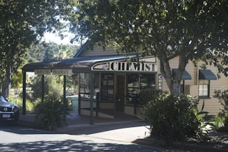 A quaint, small-town pharmacy is nestled among lush greenery with a sign reading 'chemist' prominently displayed. The building is constructed from light-colored wood and has a dark roof. A car is parked nearby, partially visible in the shadows cast by the large trees. The overall setting is serene and inviting, characteristic of a peaceful rural area.