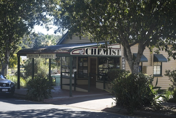A quaint, small-town pharmacy is nestled among lush greenery with a sign reading 'chemist' prominently displayed. The building is constructed from light-colored wood and has a dark roof. A car is parked nearby, partially visible in the shadows cast by the large trees. The overall setting is serene and inviting, characteristic of a peaceful rural area.