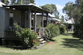 A series of rustic houses with tin roofs and wooden structures is set along a green lawn. Lush green bushes and plants with red flowers are situated near the houses, and a large wheel is placed as a decorative element near the steps of one of the houses. Trees provide a backdrop in the image, adding natural ambiance.