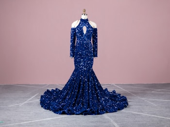 A luxurious blue sequined gown is displayed on a mannequin. The gown features a high neckline and long sleeves with cut-out shoulder details, extending into a mermaid silhouette with a dramatic train. The background is a soft pink, and the floor is light gray with subtle patterns.