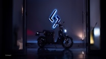 A sleek, modern-looking motorcycle is parked indoors under a dark ambiance. A bright, blue neon sign featuring a stylized symbol illuminates the scene, highlighting the bike's outline. The overall setting appears to be a showroom or an industrial space with reflective flooring.