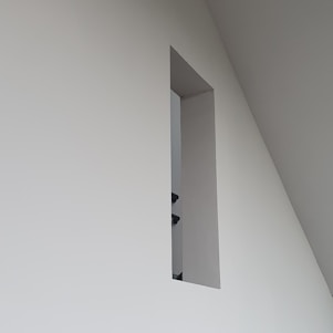 A minimalistic wall design with a rectangular cutout, revealing a glimpse of a staircase railing within a modern interior space characterized by clean lines and geometric shapes.