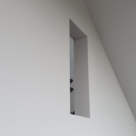A minimalistic wall design with a rectangular cutout, revealing a glimpse of a staircase railing within a modern interior space characterized by clean lines and geometric shapes.