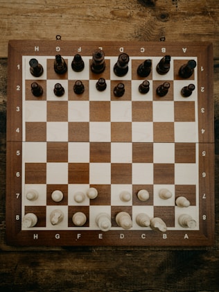 A wooden chessboard is set up with chess pieces in their starting positions. The board is placed on a rustic wooden table, and the chess pieces are made of wood, with darker pieces at the top and lighter pieces at the bottom.