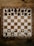A wooden chessboard is set up with chess pieces in their starting positions. The board is placed on a rustic wooden table, and the chess pieces are made of wood, with darker pieces at the top and lighter pieces at the bottom.