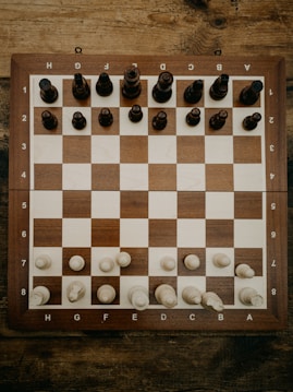 A wooden chessboard is set up with chess pieces in their starting positions. The board is placed on a rustic wooden table, and the chess pieces are made of wood, with darker pieces at the top and lighter pieces at the bottom.