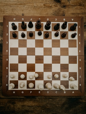 A wooden chessboard is set up with chess pieces in their starting positions. The board is placed on a rustic wooden table, and the chess pieces are made of wood, with darker pieces at the top and lighter pieces at the bottom.
