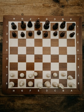 A wooden chessboard is set up with chess pieces in their starting positions. The board is placed on a rustic wooden table, and the chess pieces are made of wood, with darker pieces at the top and lighter pieces at the bottom.